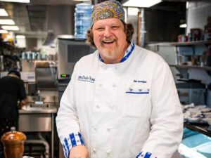 Smiling Bill Ranniger wearing a colorful beanie and Duke's Seafood chef robe