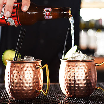 Bottle of beer pouring into one of two copper Moscow Mule mugs